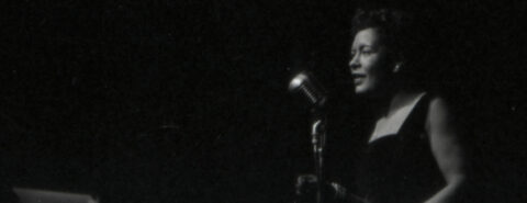 Black and white image of jazz singer Billie Holiday standing in front of a mic