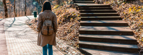 Woman in brown coat and blue denim jeans , wearing a backpack walking on brick path during daytime