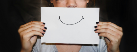 A sad woman holding a piece of paper with a smile drawn on it in front of her mouth